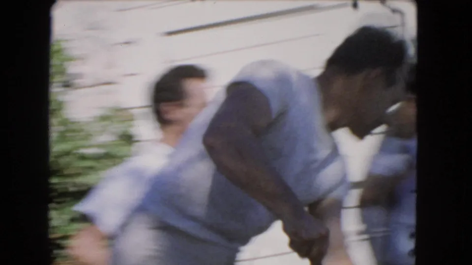 Motion blur shows adult men moving close together in a 1966 Los Angeles residential outdoor scene near a pale wall and greenery.
