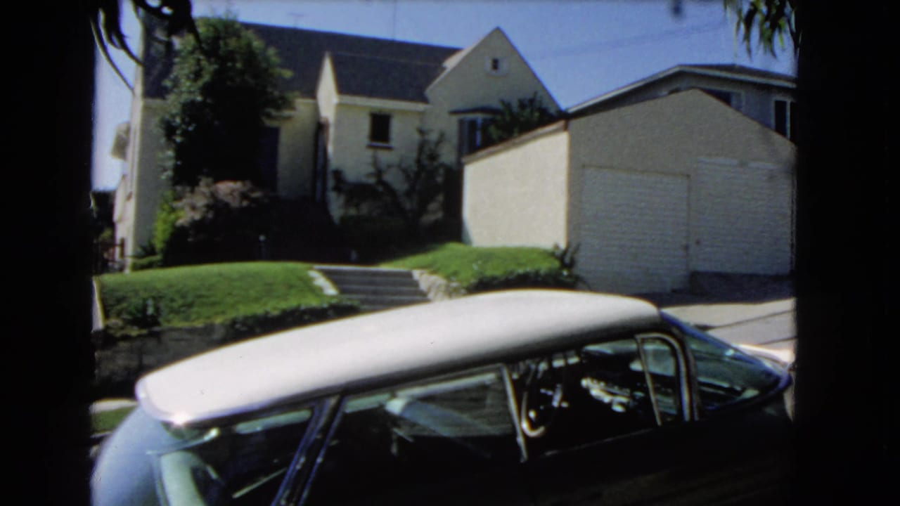 1966 Los Angeles home-movie frame showing a dark car with a pale roof parked across from a light-colored house, garage, lawn, and trees.