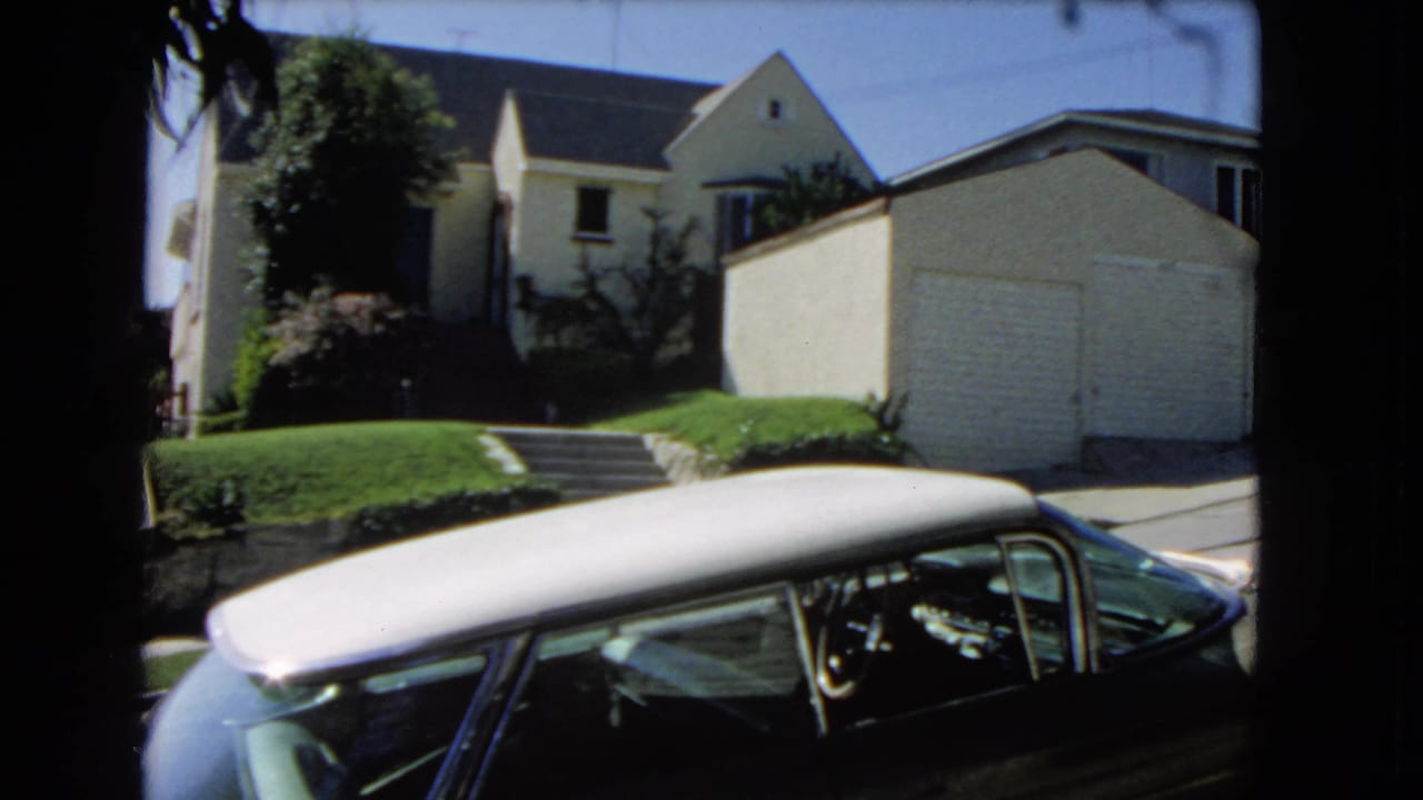 Parked car in the foreground with a light house, garage, lawn, and trees behind it.