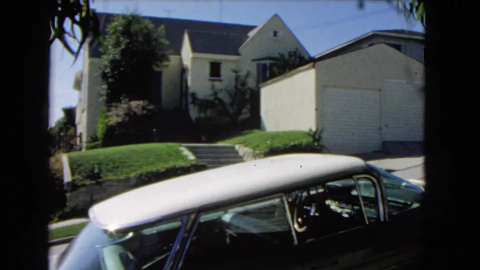 1966 Los Angeles home-movie view of a light house and detached garage above a lawn, with a dark parked car partly filling the foreground.