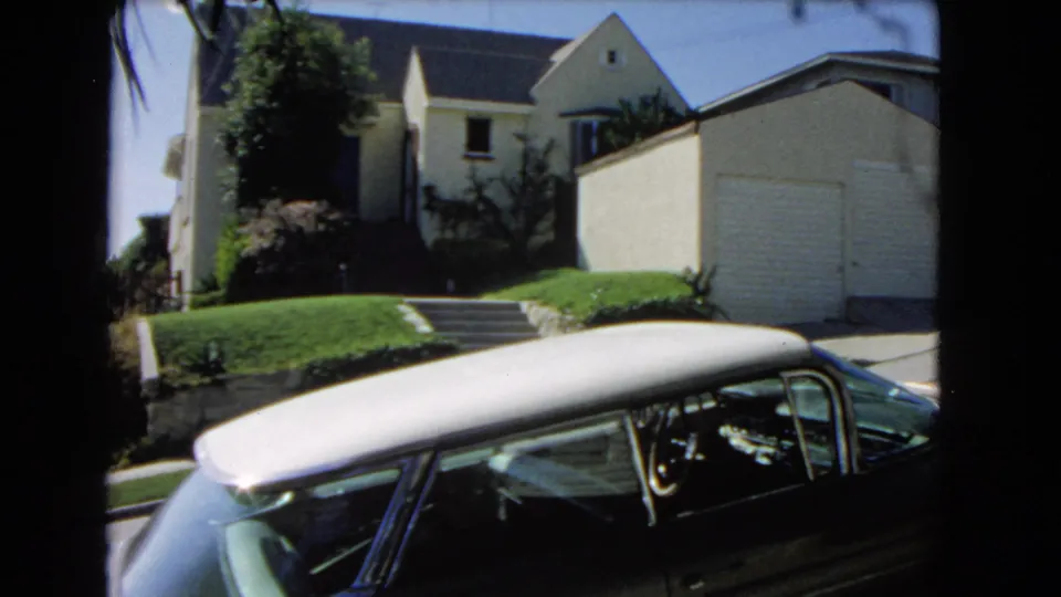 1966 Los Angeles suburban street frame with a dark car in the foreground and a light-colored house, garage, lawn, steps, and trees behind it.