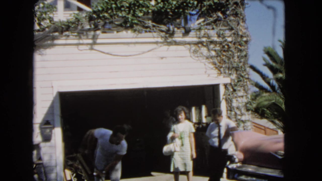 1966 Los Angeles driveway scene with a white couple near an open garage and a Black man bending near the ground.