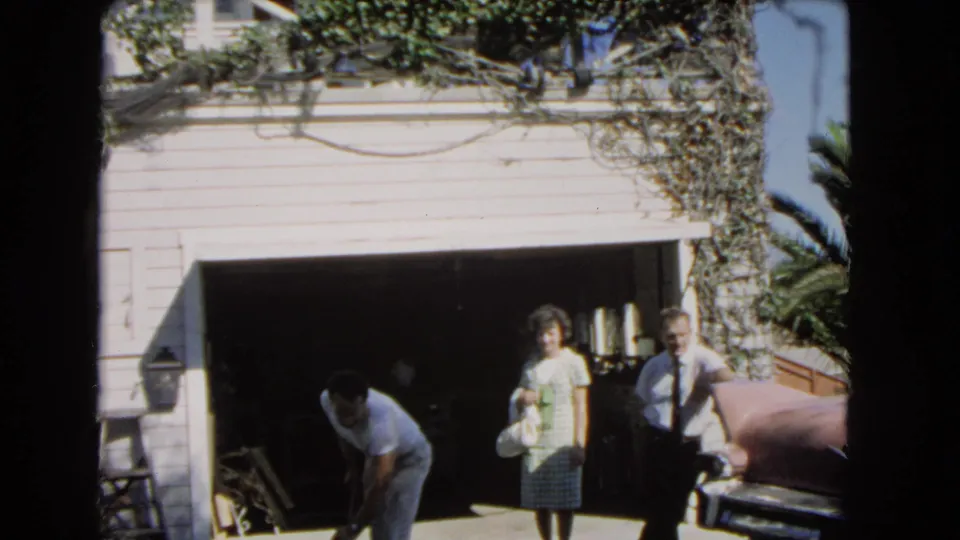 1966 Los Angeles home-movie frame of a couple standing by an open garage while a Black man works near the driveway.