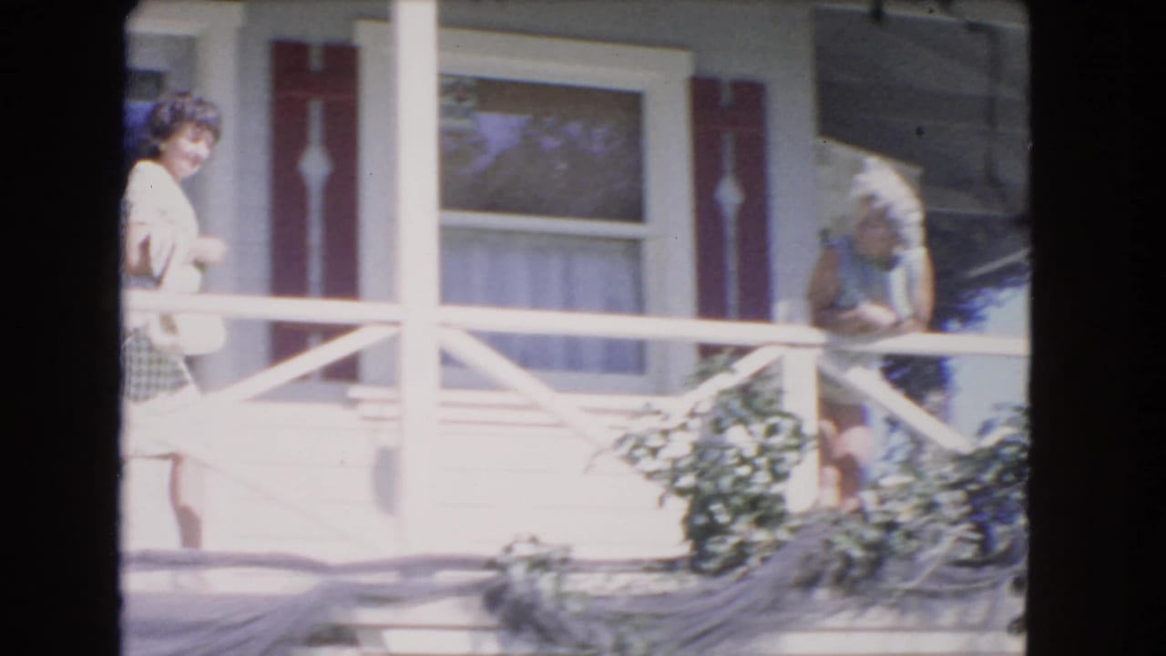 Young woman walks along a porch while an older woman sits or leans nearby.