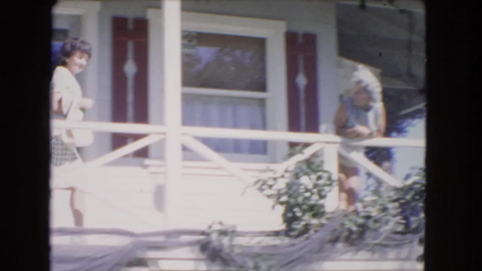 A young woman moves along the Los Angeles porch as an older woman rests near the railing, with foliage and house windows visible.
