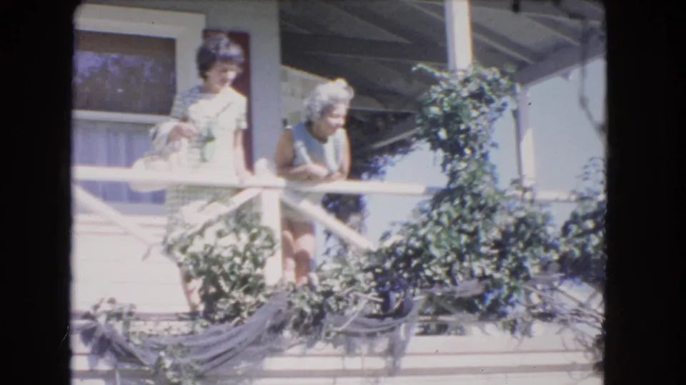 The younger woman stands near the older woman on the Los Angeles porch, partly framed by leafy vines and the white railing.