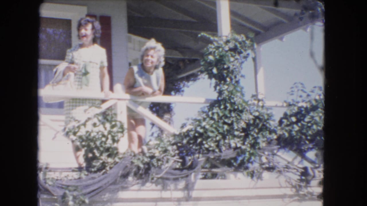 Two women stand on a sunlit porch behind a white railing and vines.