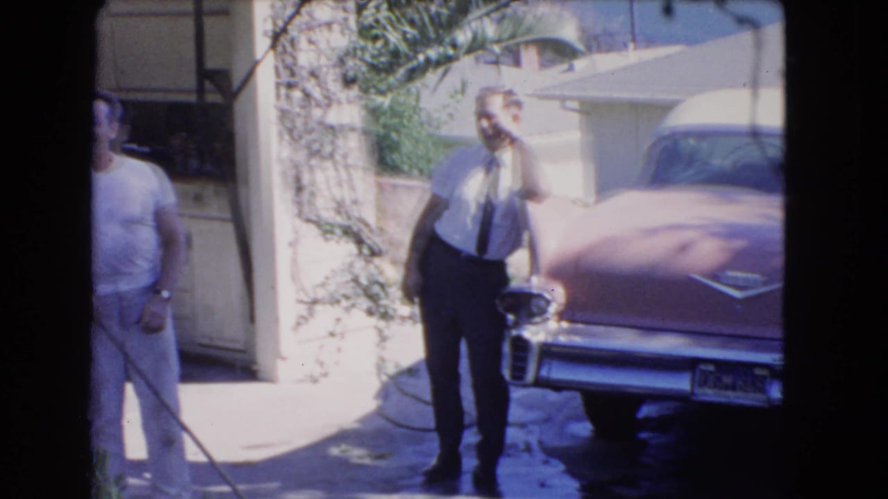 A man in a shirt and tie stands beside a pink car near a driveway.