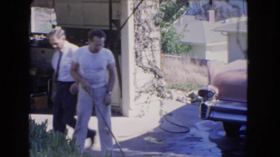 Two men talk near a driveway and partially visible pink car in 1966 Los Angeles home-movie footage.