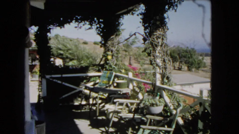 A shaded Los Angeles porch or patio view shows railings, plants, outdoor furniture, and sunlit hillside beyond.