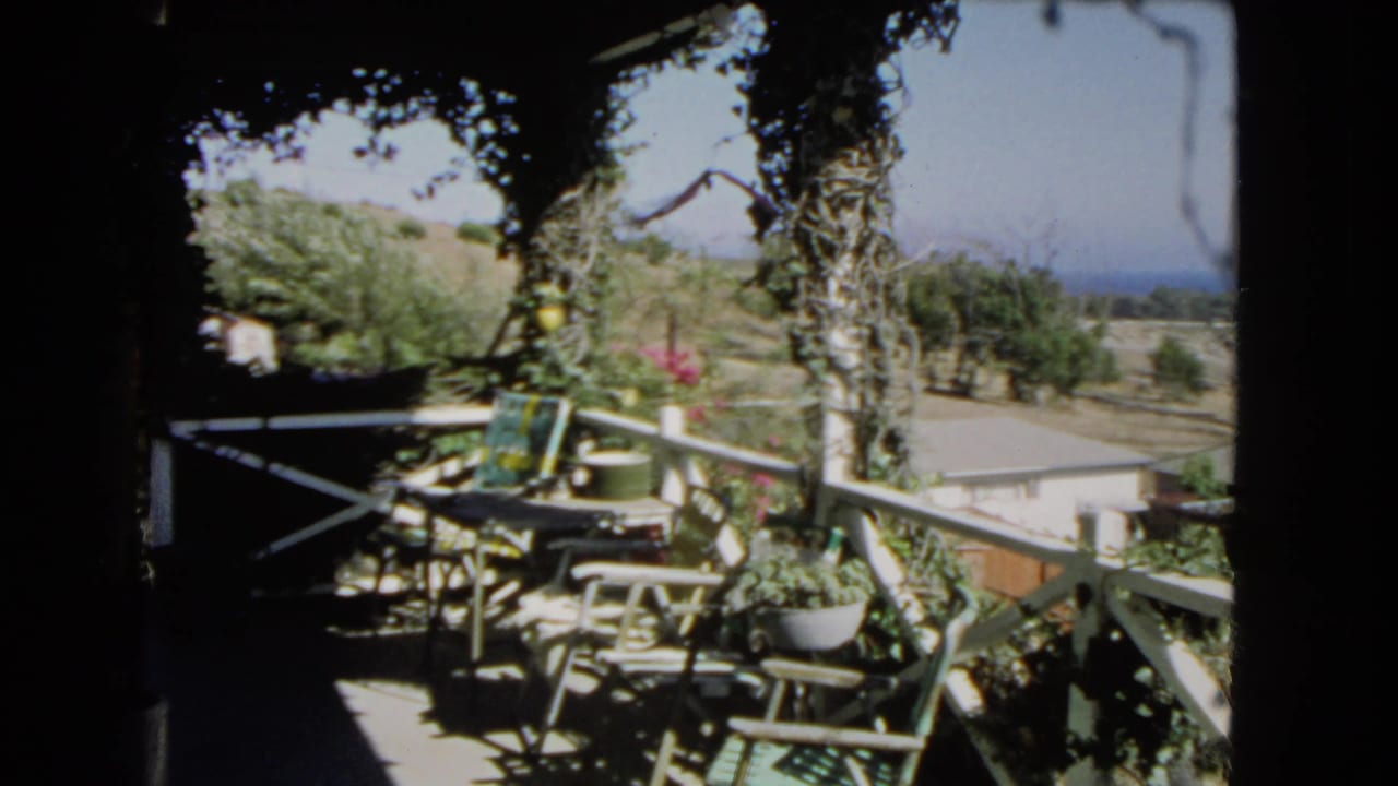 Shaded balcony with patio chairs, potted plants, white railing, trees, hillside, and distant ocean.