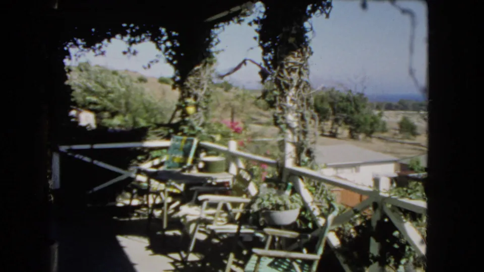 1966 Los Angeles home-movie frame from a shaded balcony, showing patio furniture, vines, potted plants, hillside trees, and distant sea.