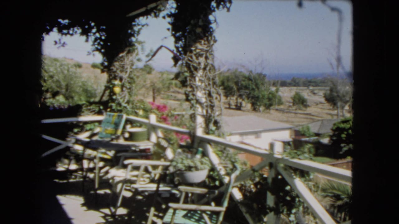 Balcony railing and patio furniture under shade, with garden plants, trees, hillside, and ocean beyond.