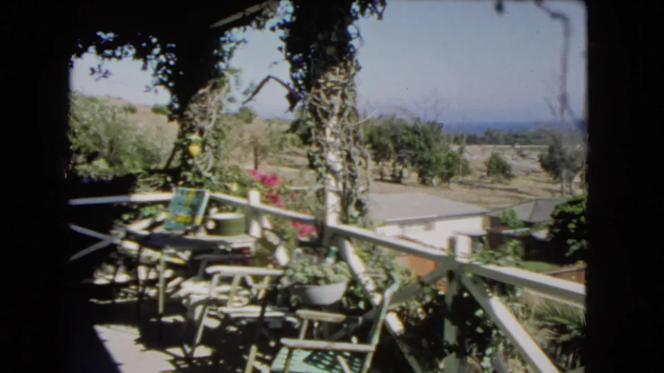 Shaded 1966 Los Angeles balcony view with white railings, chairs, table, flowering plants, hillside trees, and blue ocean in the distance.