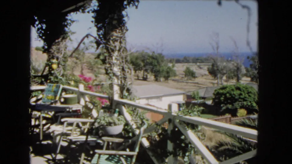 Home-movie frame from a Los Angeles balcony in 1966, with patio chairs, potted plants, roofline below, hillside trees, and distant ocean.