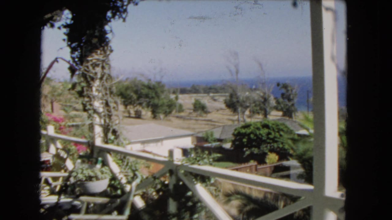 Balcony railing frames a sunny coastal neighborhood with rooftops, palms, and ocean beyond.
