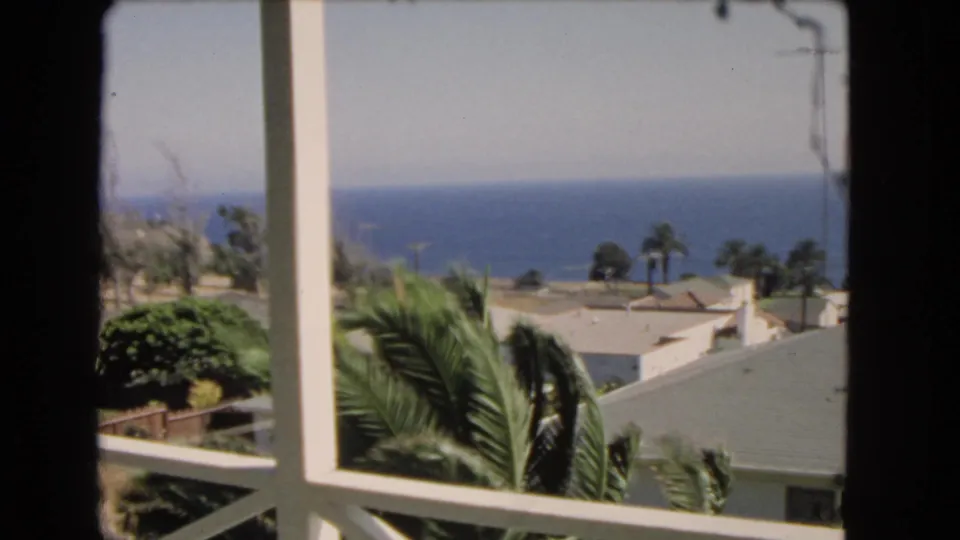 1966 coastal Los Angeles-area home-movie frame showing balcony railings, palms, nearby rooftops, and the Pacific Ocean under clear light.