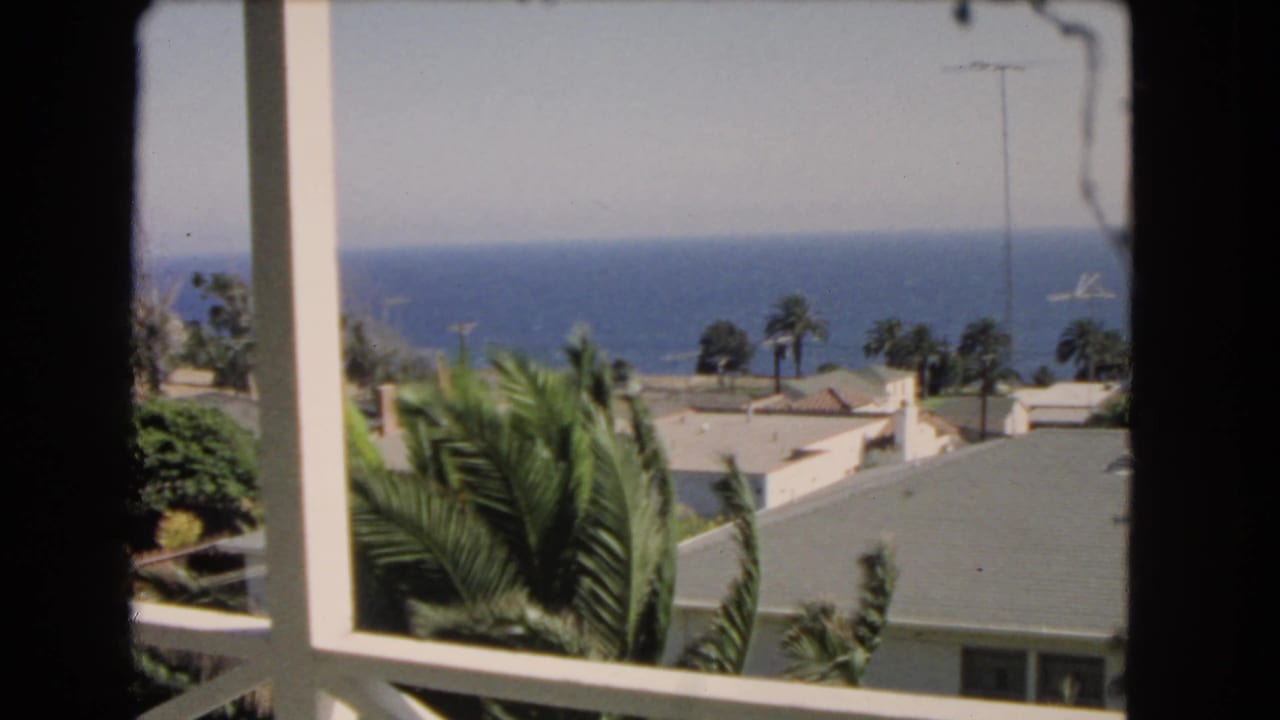 1966 Los Angeles home-movie frame showing a balcony view with palm trees, rooftops, ocean, and a distant vessel.