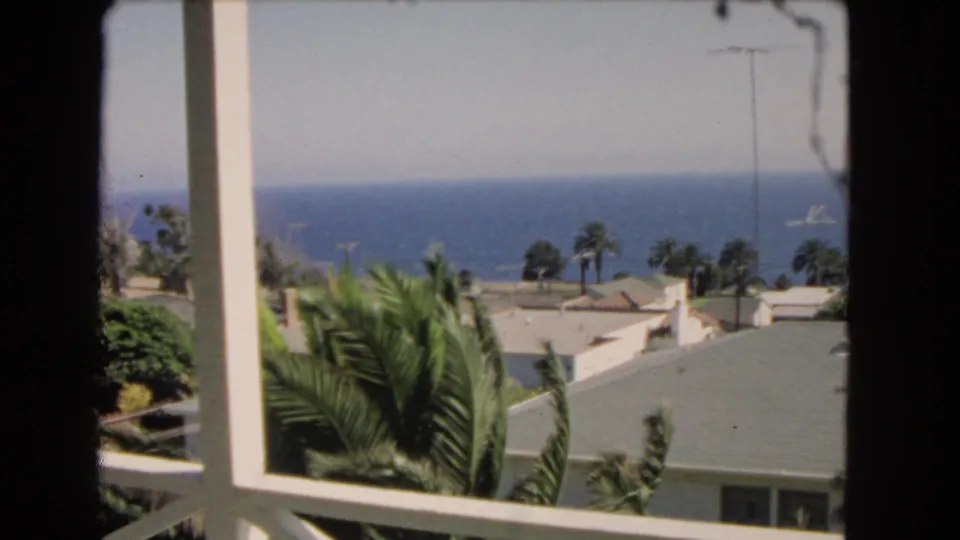 1966 Los Angeles home-movie frame showing a balcony view with palm trees, rooftops, ocean, and a distant vessel.