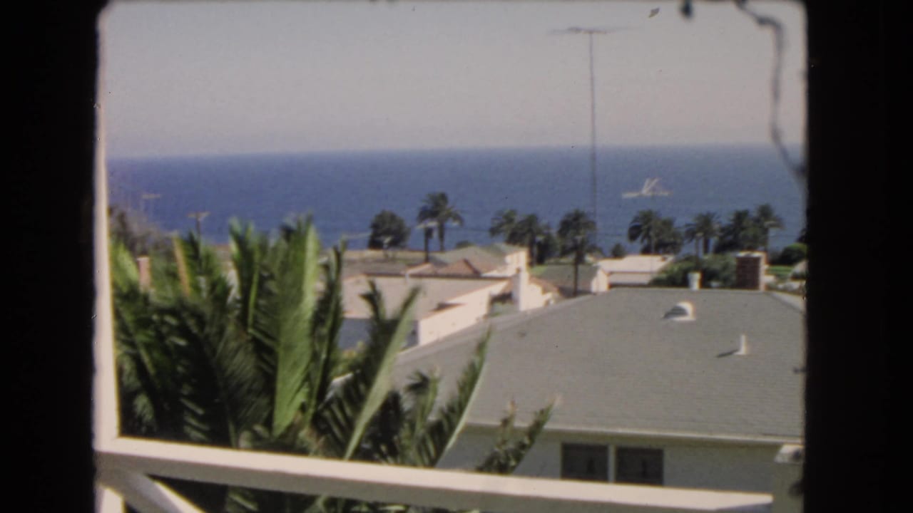 1966 Los Angeles coastal view from a balcony, with palms, pale rooftops, blue water, and a small vessel offshore.