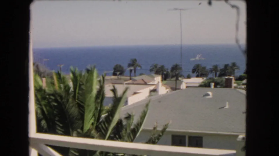 1966 Los Angeles coastal view from a balcony, with palms, pale rooftops, blue water, and a small vessel offshore.