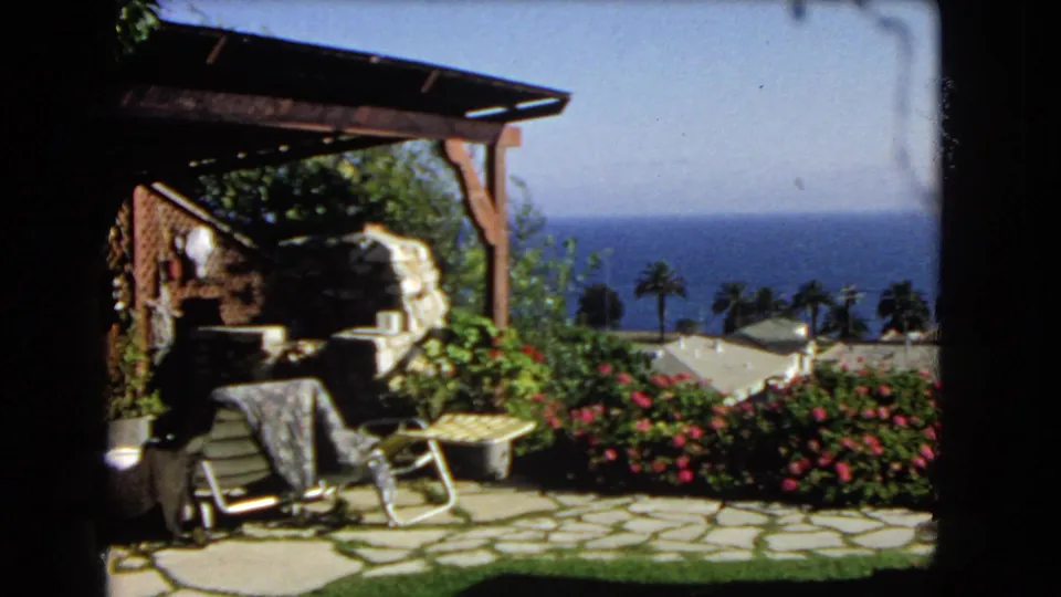 1966 Los Angeles coastal patio scene with lounge chairs, stone paving, flowers, palms, rooftops, and ocean horizon.
