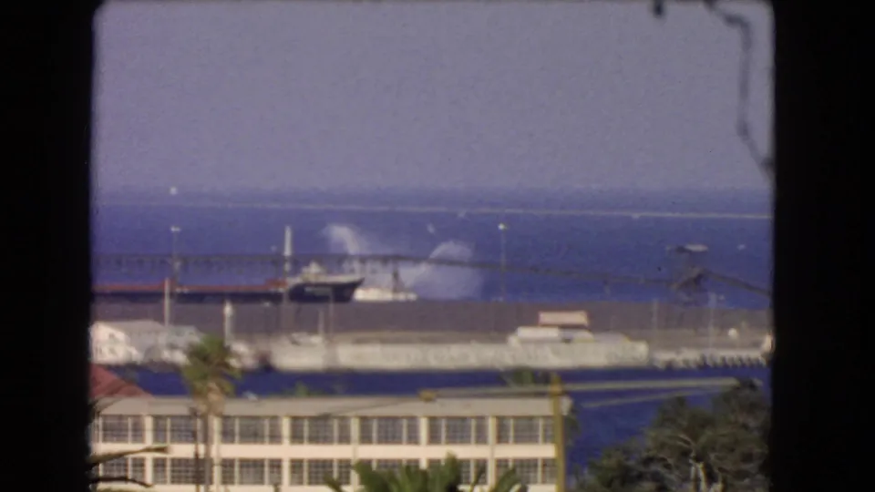 A distant Los Angeles waterfront or harbor view shows low buildings, water, and a hazy shoreline in 8mm color.