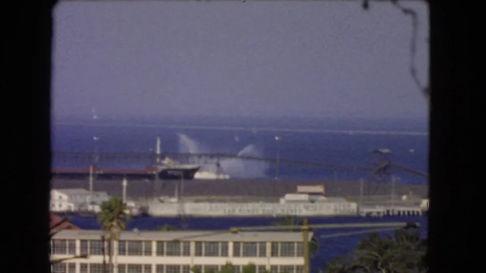 1966 Los Angeles harbor scene showing a docked vessel spraying water near a pier, with foreground buildings and palms.