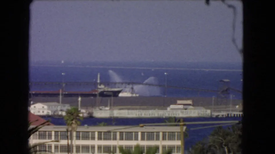 1966 Los Angeles waterfront view with a vessel spraying water near pier structures under a hazy sky.