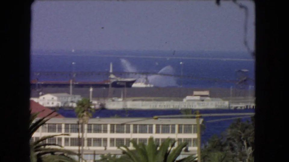 1966 Los Angeles harbor footage frame showing a docked ship spraying water beyond foreground buildings and palm fronds.