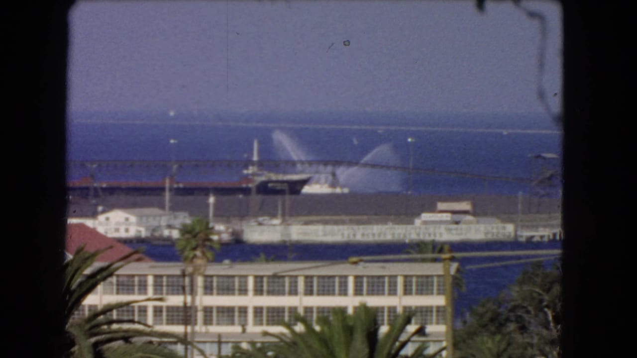 1966 Los Angeles harbor scene framed by a window edge, showing palms, waterfront structures, a pier, and a vessel spraying water offshore.