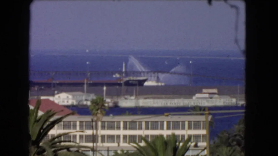 1966 Los Angeles harbor view with palms and waterfront buildings in the foreground and a vessel spraying water near the pier under a hazy sky.