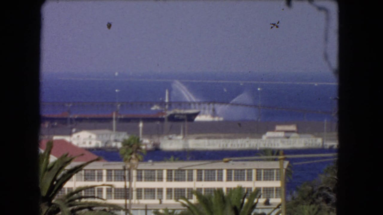1966 Los Angeles coastal harbor view with palm trees, waterfront buildings, blue ocean, and a distant vessel spraying water near a pier.