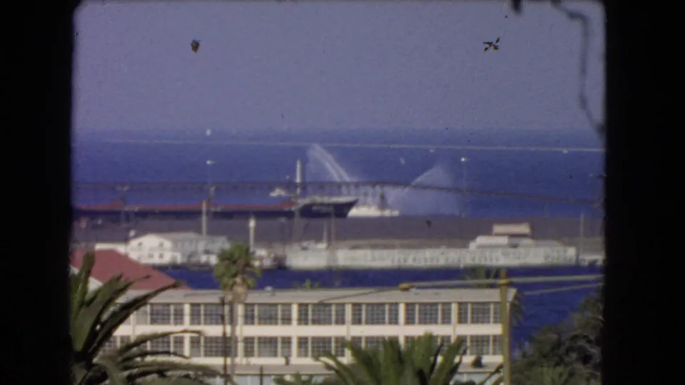 1966 Los Angeles coastal harbor view with palm trees, waterfront buildings, blue ocean, and a distant vessel spraying water near a pier.