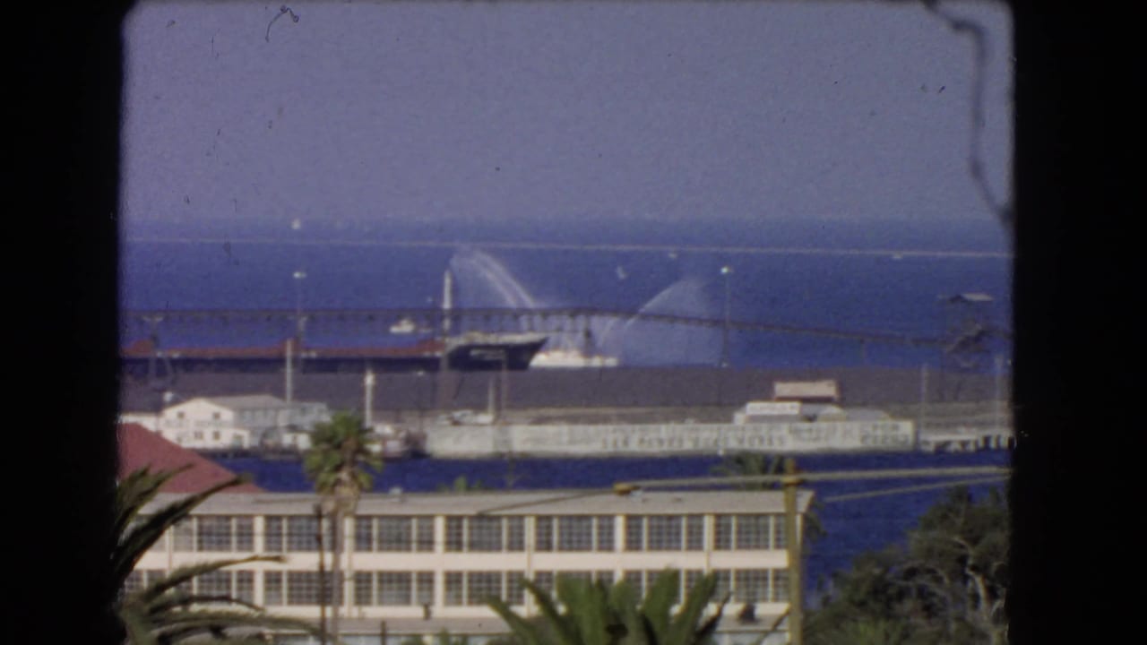 1966 Los Angeles waterfront seen through dark foreground framing, with palm trees, harbor buildings, ocean, and a distant vessel spraying water.