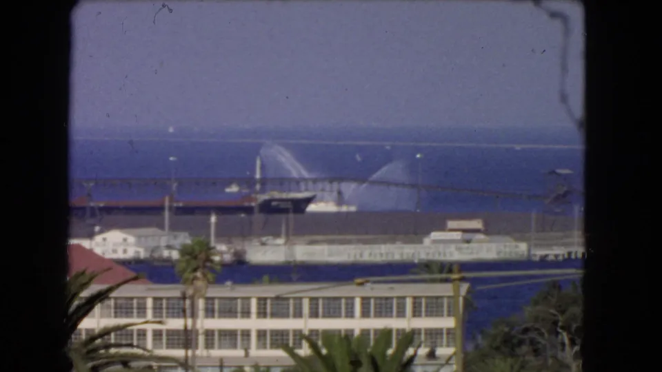 1966 Los Angeles waterfront seen through dark foreground framing, with palm trees, harbor buildings, ocean, and a distant vessel spraying water.
