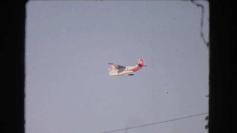 A small red-and-white seaplane crosses pale sky, with a faint diagonal line visible near the lower part of the 1966 Los Angeles frame.