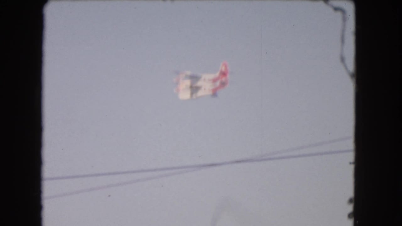 1966 Los Angeles 8mm frame showing a small red and white airplane crossing a pale sky above utility wires.