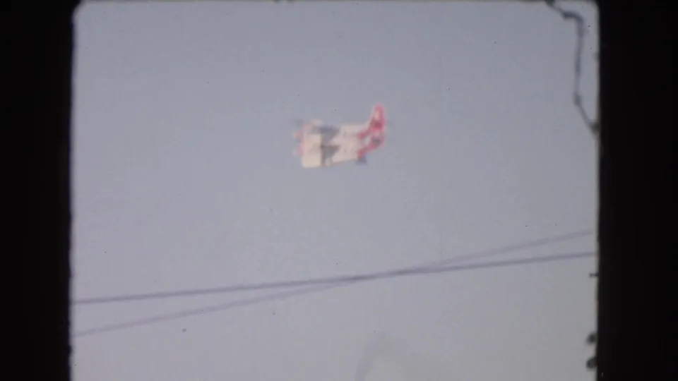 1966 Los Angeles 8mm frame showing a small red and white airplane crossing a pale sky above utility wires.