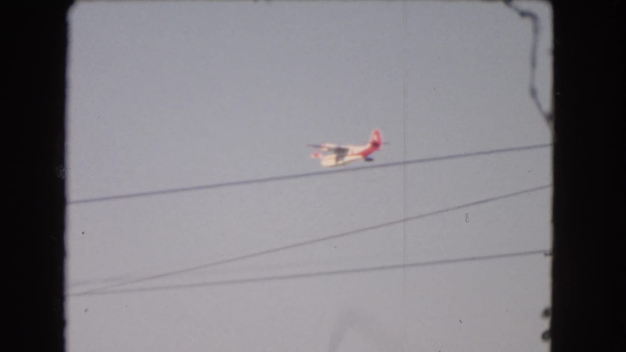 1966 Los Angeles home-movie frame with a small airplane moving near overhead utility lines in a pale blue sky.