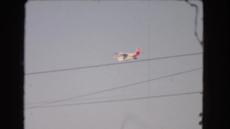 1966 Los Angeles home-movie frame with a small airplane moving near overhead utility lines in a pale blue sky.