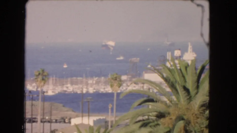 1966 Los Angeles coastal frame showing palms, marina masts, water, distant vessels, and a small airplane above the shoreline haze.