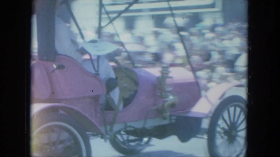 Pink antique-style car with a driver and passenger passes blurred spectators in 1966 Milwaukee home-movie footage.