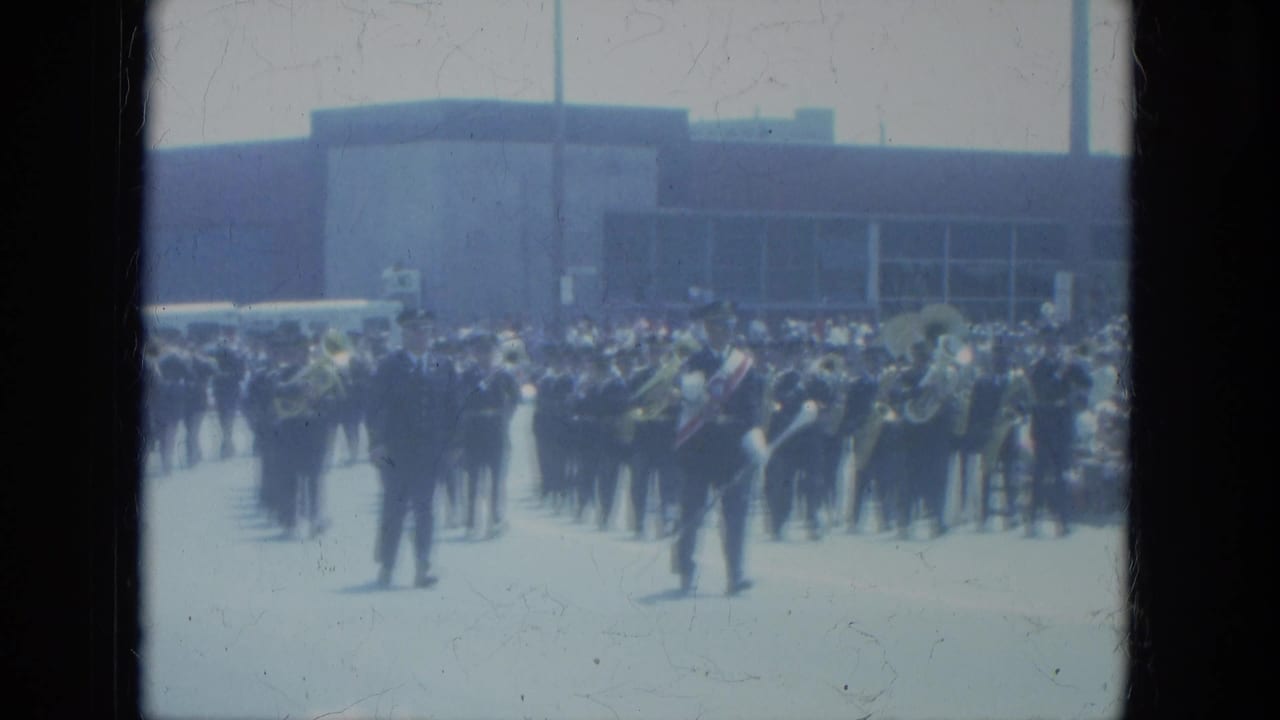 1966 Milwaukee home-movie frame showing a marching band moving along a city street with spectators in the background.