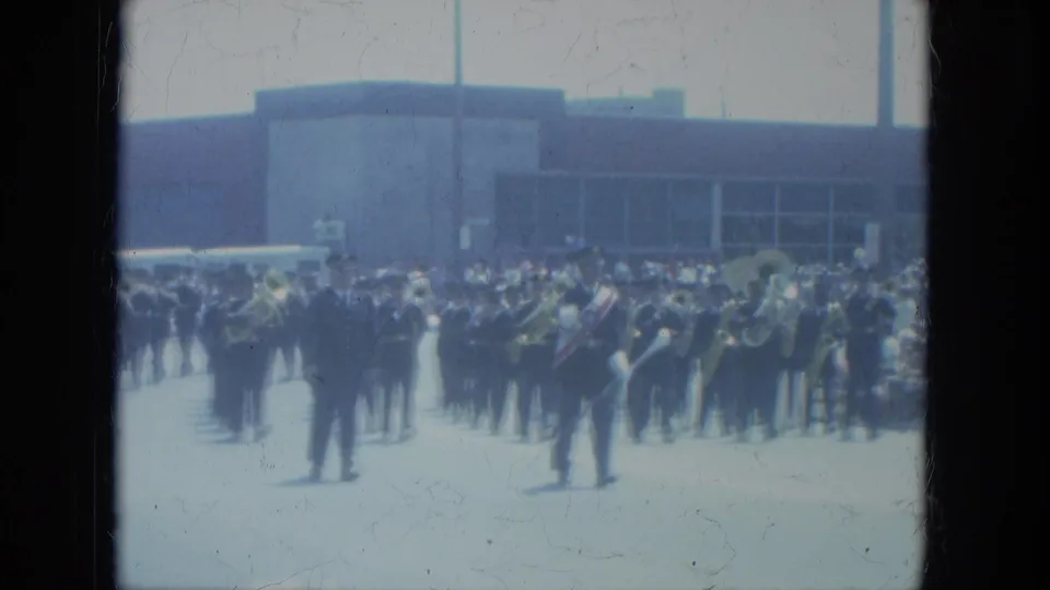 1966 Milwaukee home-movie frame showing a marching band moving along a city street with spectators in the background.
