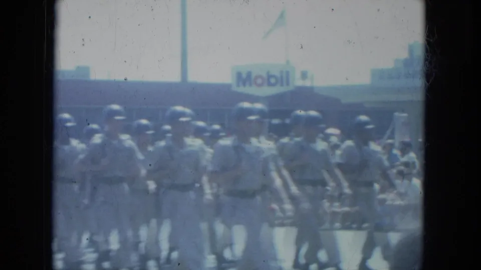 Uniformed soldiers march in formation on a Milwaukee street, with a Mobil gas station sign visible behind them.