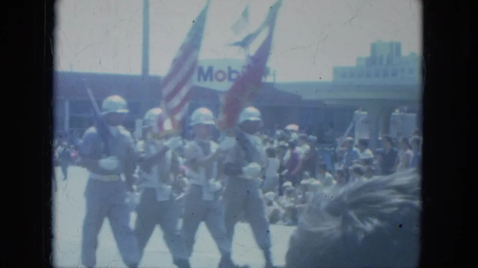 Soldiers carrying U.S. flags and rifles pass spectators on a Milwaukee street, with the Mobil sign in the background.