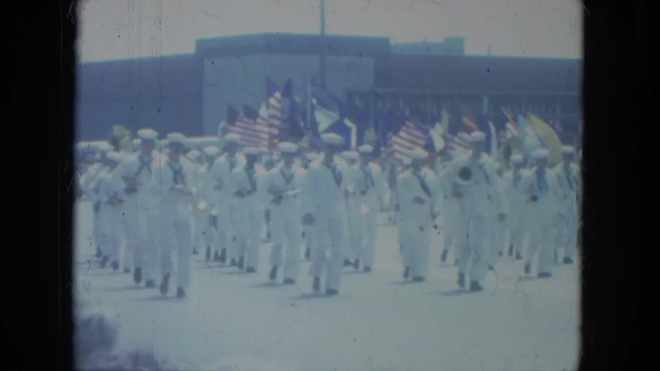 1966 Milwaukee home-movie frame showing sailors in white uniforms marching with American flags in a civic parade.