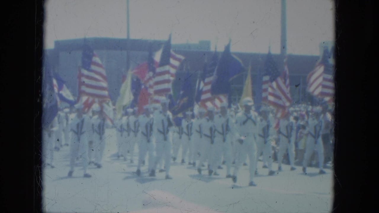 Flag-bearing parade group marches in front of a crowd and low building.