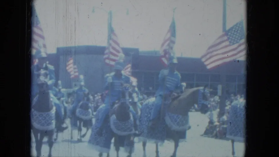1966 Milwaukee parade view with horses wearing patterned coverings while riders and American flags fill the street scene.